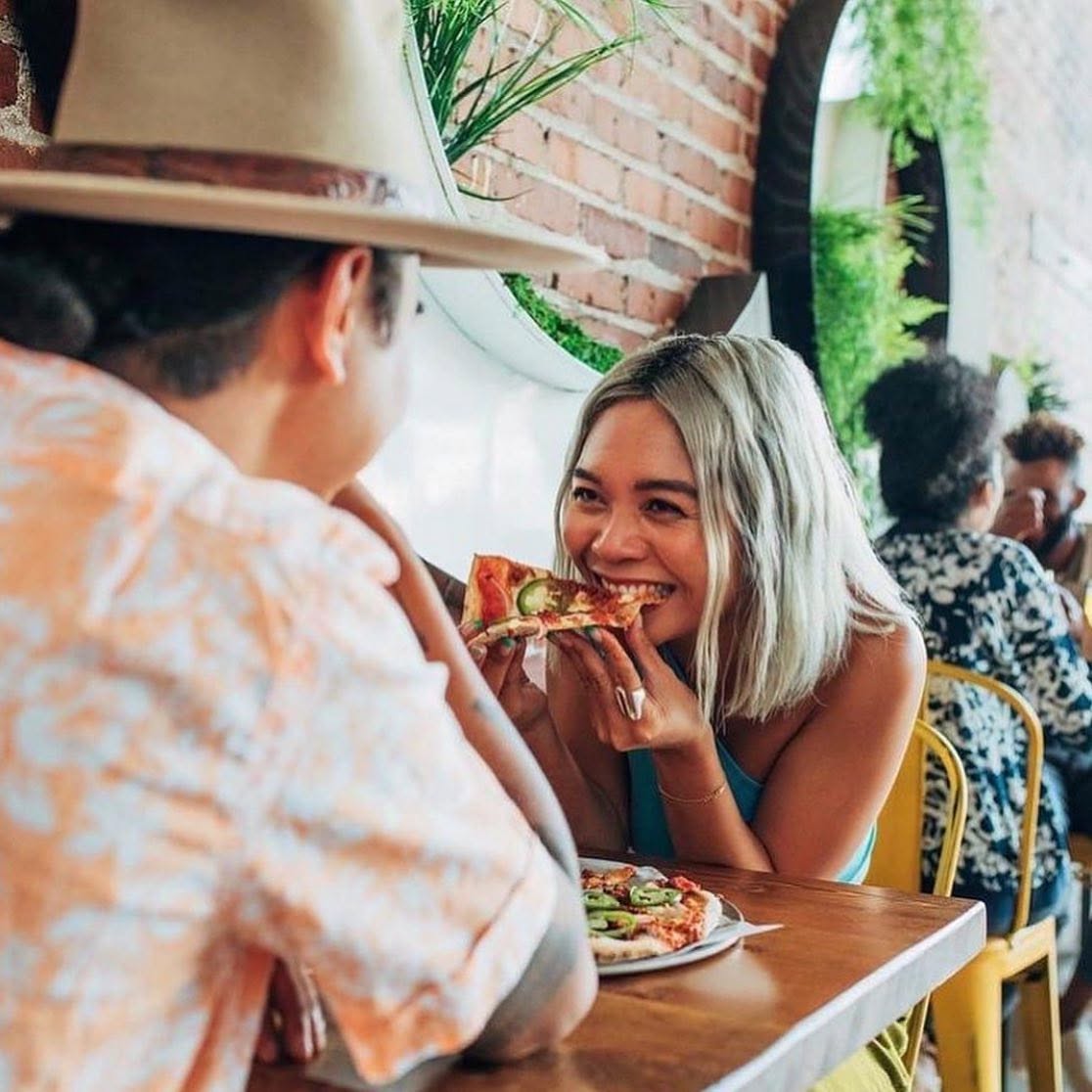 A women eating a slice of pizza while sitting across from a man in a hat